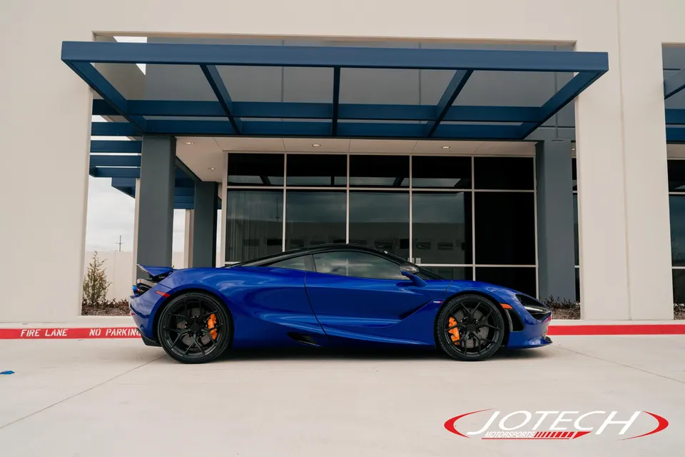 A blue McLaren 720S supercar with black wheels and orange brake calipers parked in front of a modern building.