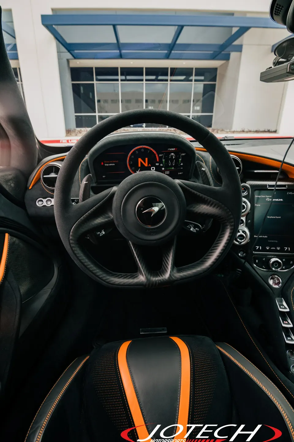 Close-up view of a McLaren's interior, featuring the carbon fiber steering wheel, digital dashboard, and black and orange seats.