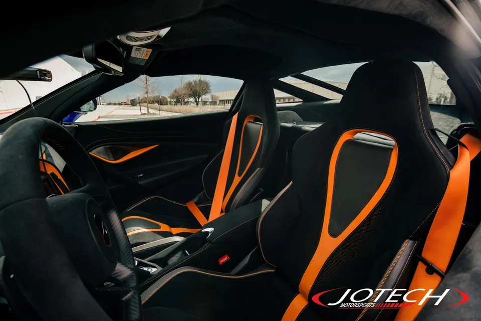 Interior view of a McLaren with black and orange racing seats, orange harnesses, and a carbon fiber steering wheel.