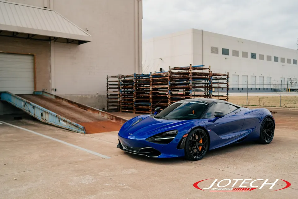 A blue McLaren 720S supercar parked in an industrial area with a loading dock and warehouse in the background.