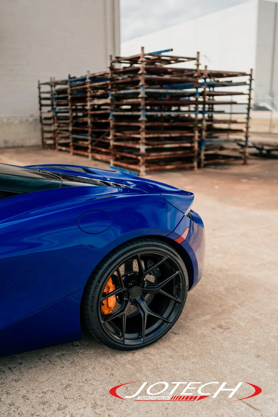 Blue McLaren with black aftermarket wheels and orange brake calipers parked in an industrial setting.