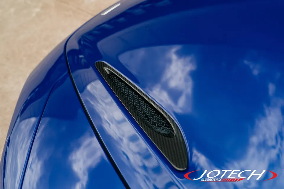 Close-up of a blue car's hood with a carbon fiber vent and honeycomb mesh.