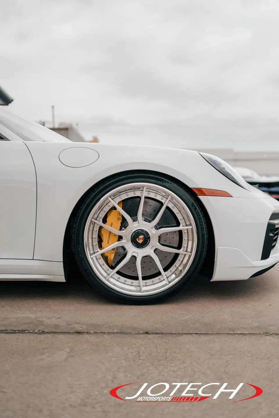 Close-up of a white Porsche's front wheel with custom silver multi-spoke wheels and yellow brake calipers.