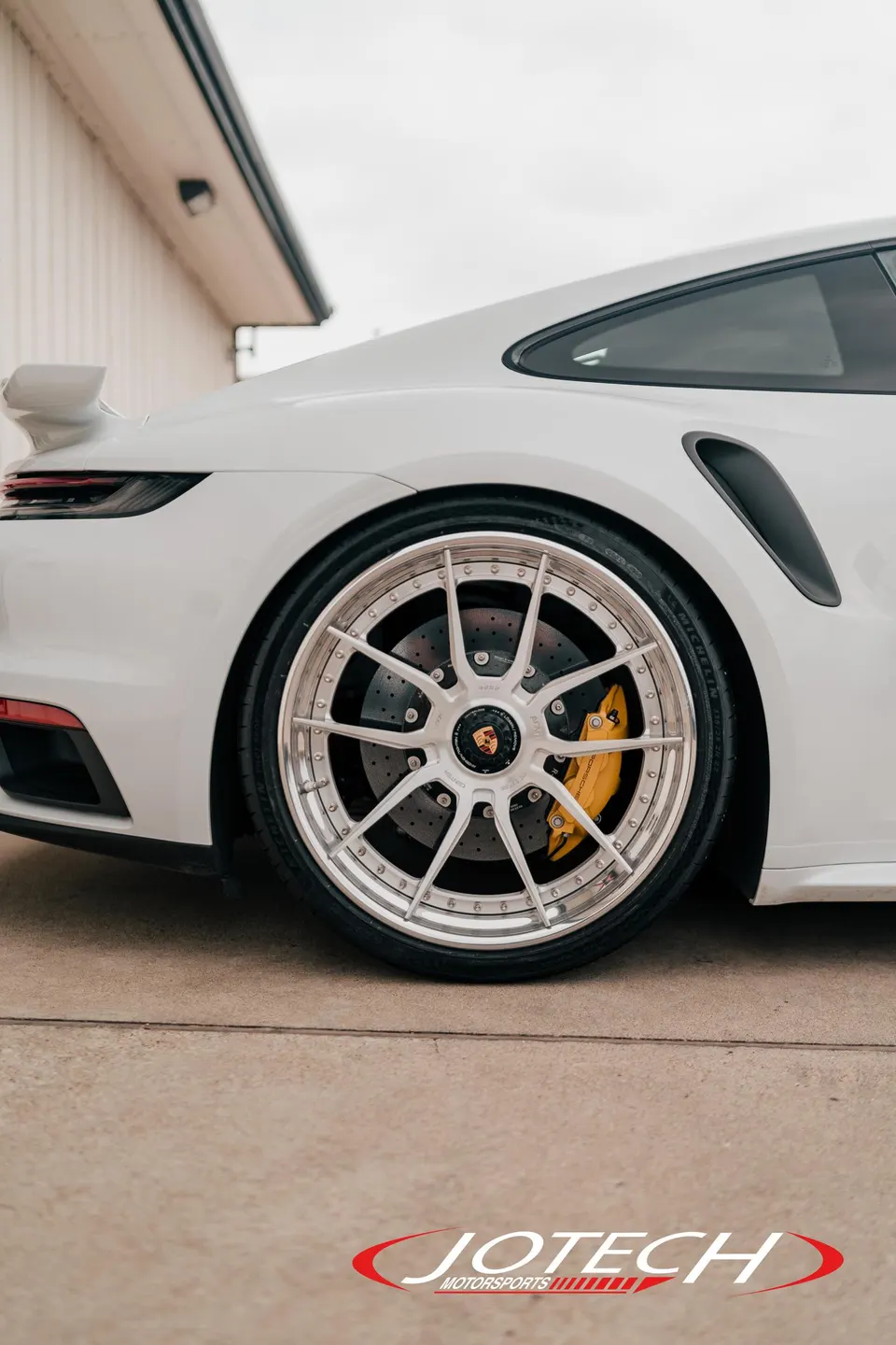 A close-up of the rear wheel of a white Porsche 911, featuring multi-spoke silver wheels and yellow brake calipers.