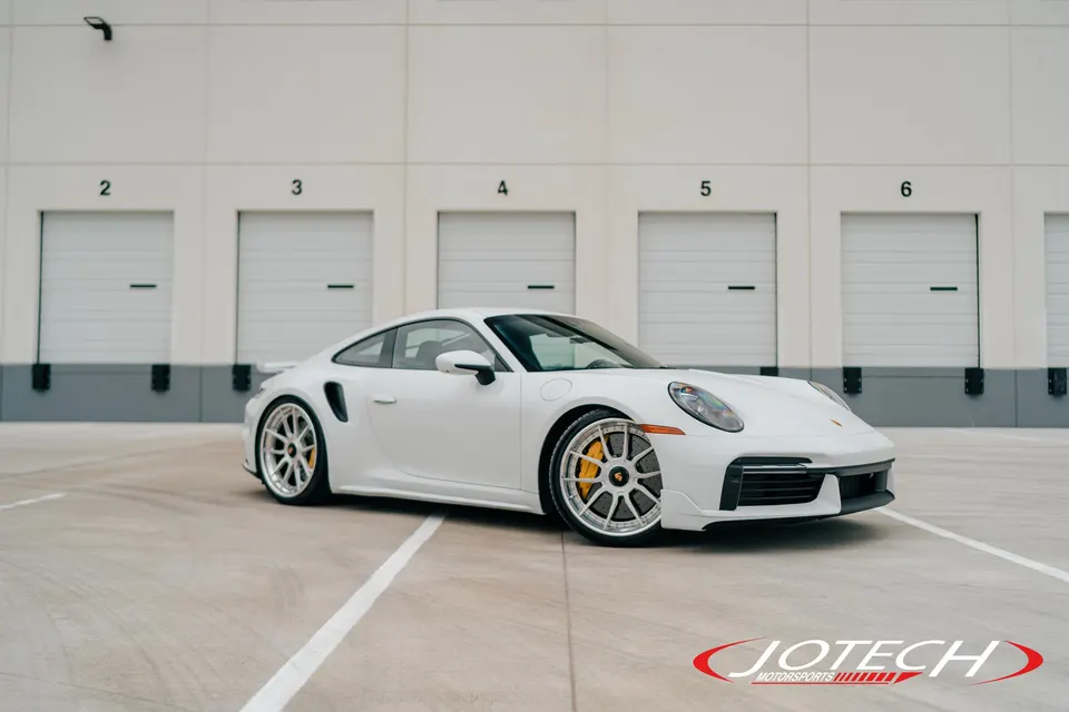 White Porsche 911 Turbo S with aftermarket multi-spoke wheels and yellow brake calipers parked in front of a warehouse.