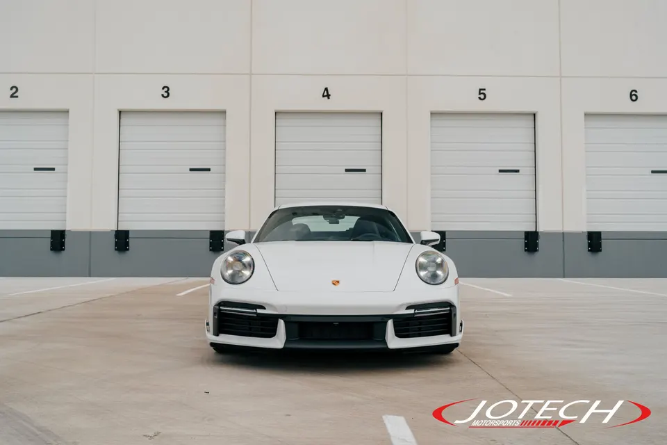 A white Porsche 911 parked in front of a warehouse with multiple loading docks, showcasing its front design.