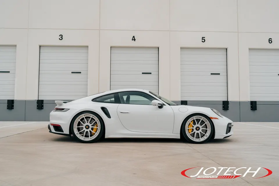 White Porsche 911 Turbo S with aftermarket multi-spoke wheels and yellow brake calipers parked in front of garage doors.