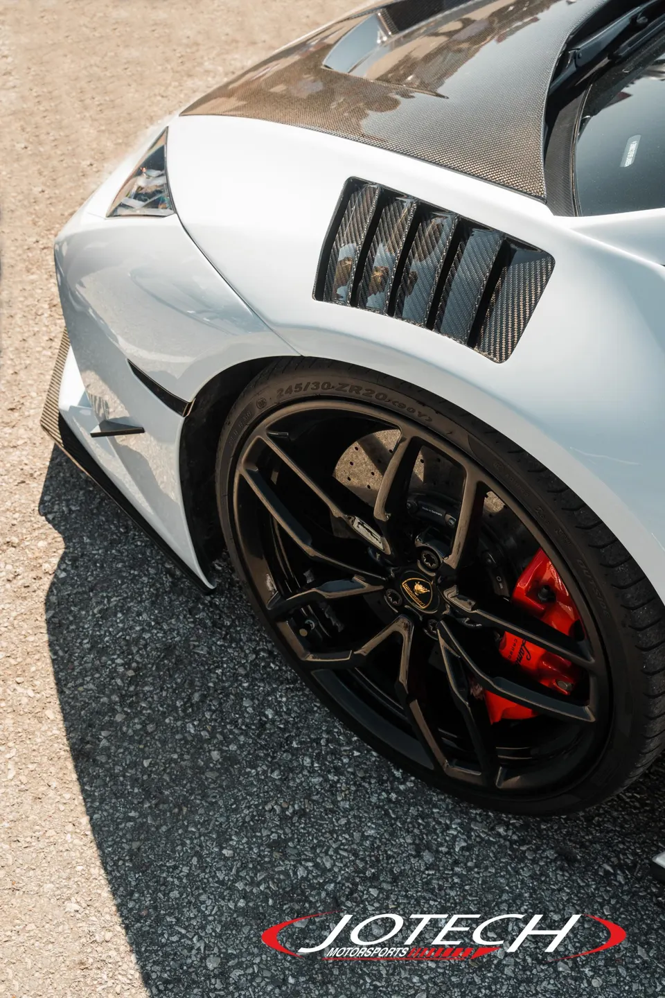 A close-up of a white Lamborghini's front wheel, featuring black multi-spoke rims, red brake calipers, and carbon fiber fender vents.