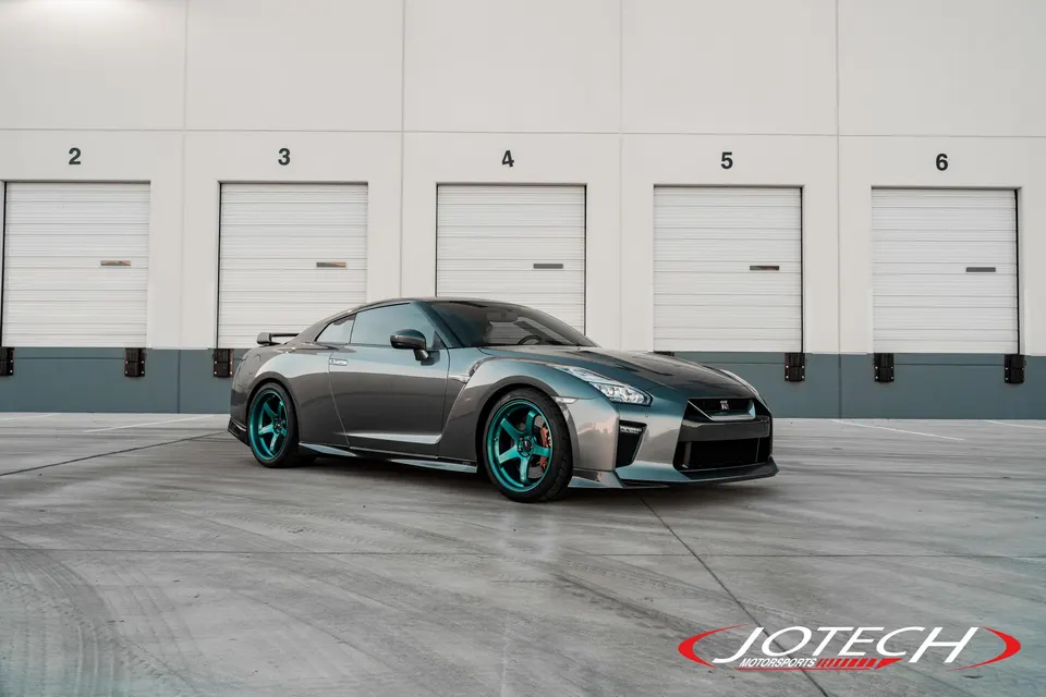 Grey Nissan GT-R with teal wheels parked in front of a warehouse, showcasing its custom look.