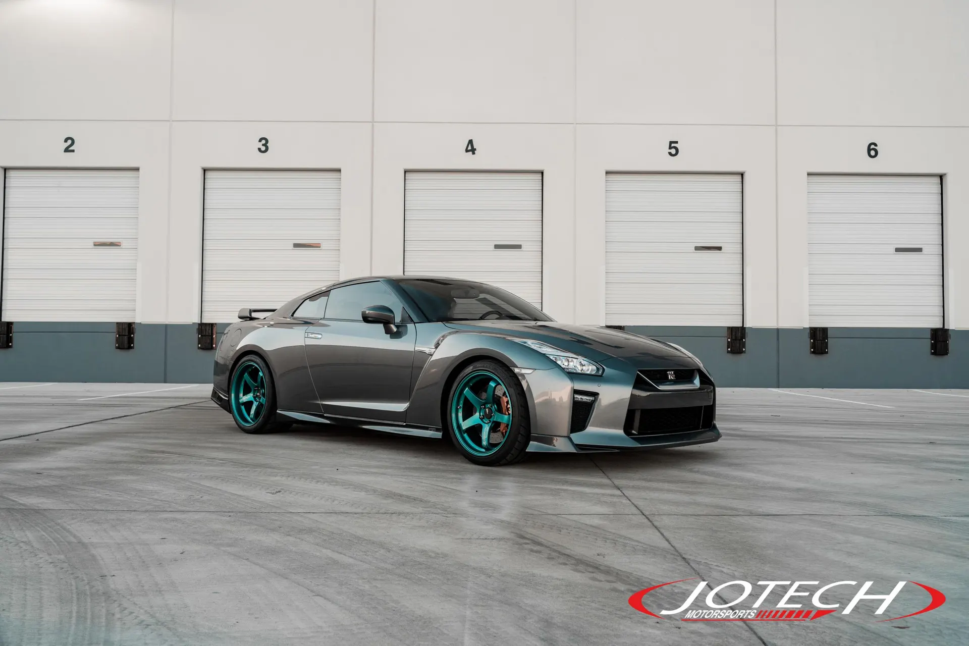 Grey Nissan GT-R with teal wheels parked in front of a warehouse, showcasing its custom look.