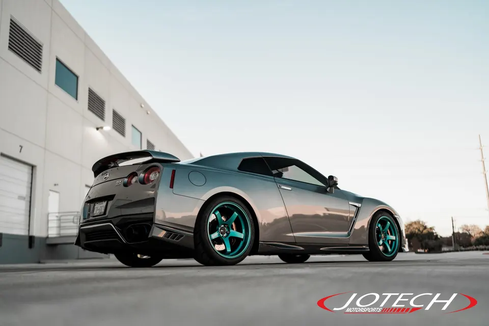 A silver Nissan GT-R with teal aftermarket wheels and a rear spoiler parked in front of a modern building.
