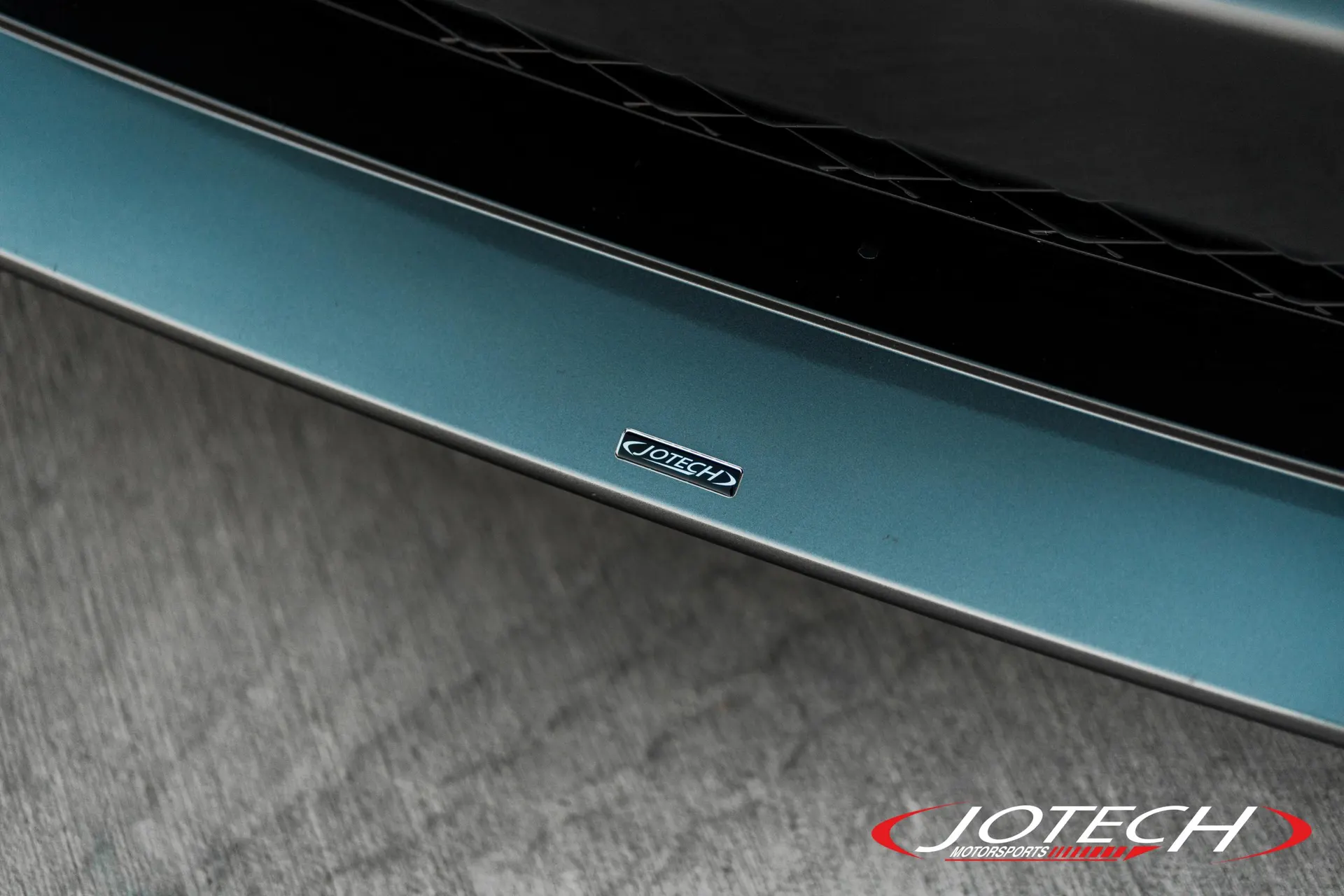 Close-up of a Jotech badge on a teal front splitter of a performance car, showcasing exterior detail.