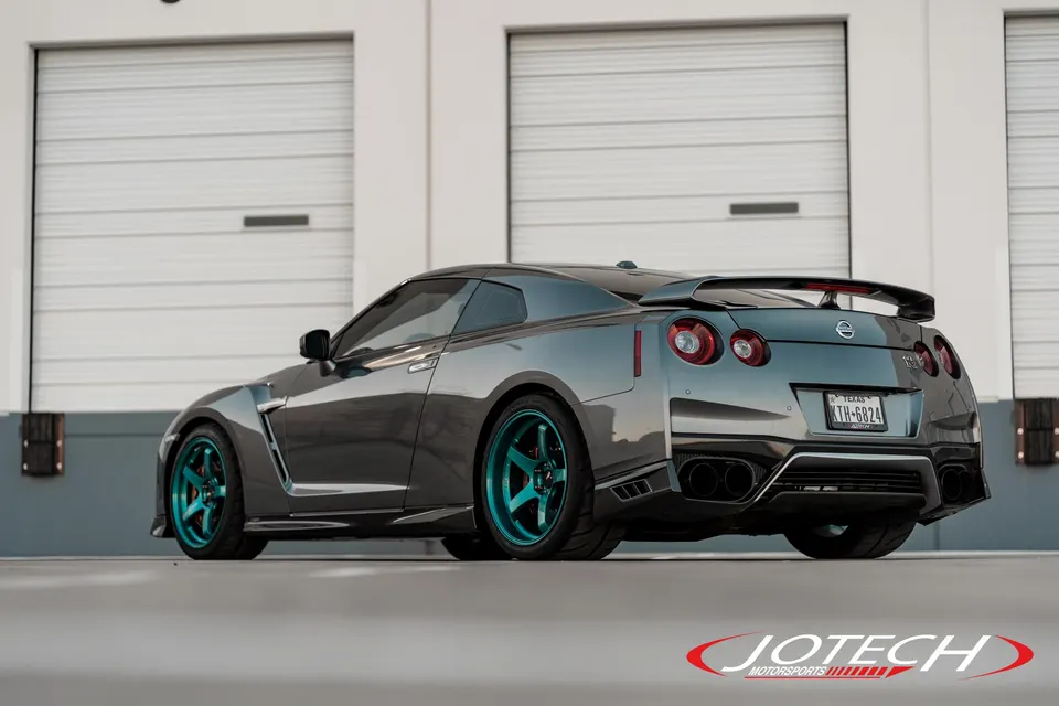 A grey Nissan GT-R with striking teal aftermarket wheels and a custom rear wing parked in front of white garage doors.