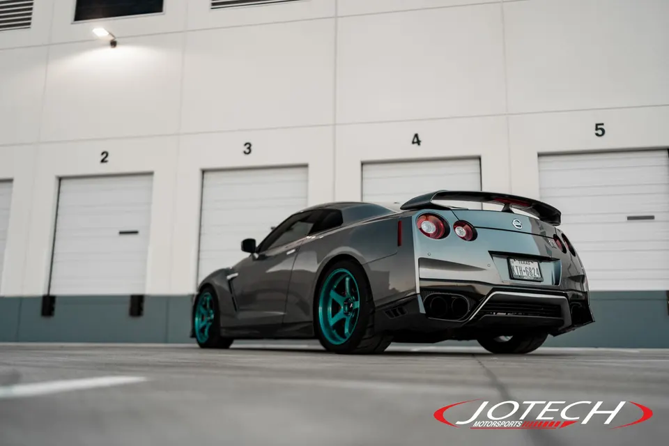 Rear view of a grey Nissan GT-R with custom teal aftermarket wheels parked in front of a warehouse.