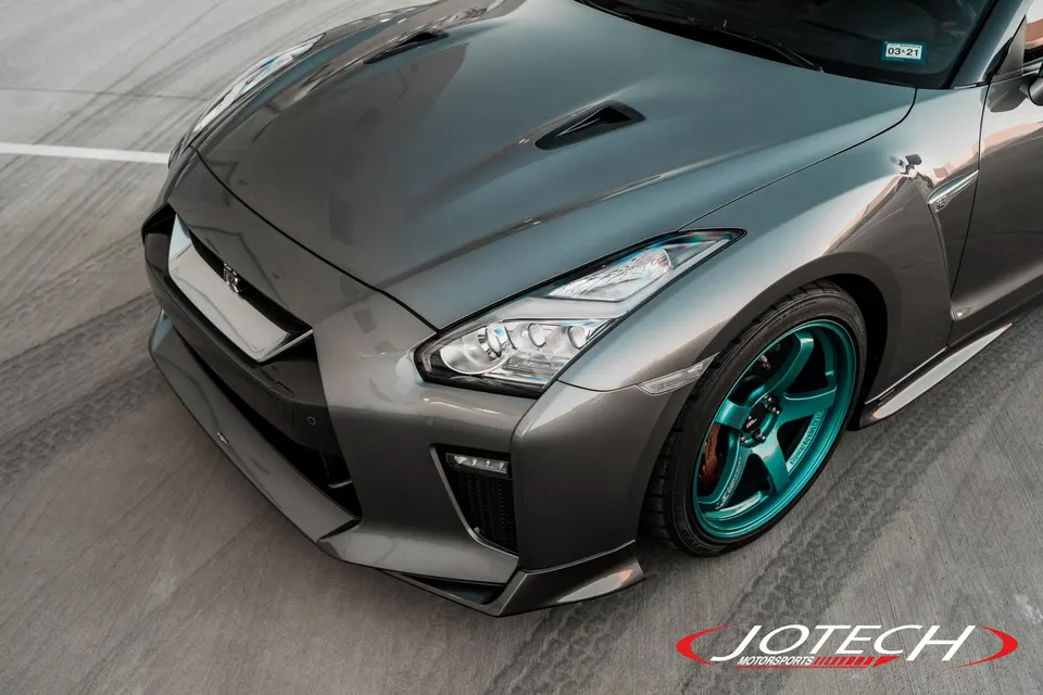 A grey Nissan GT-R R35 with aftermarket front bumper, splitter, and teal wheels parked on a concrete surface.