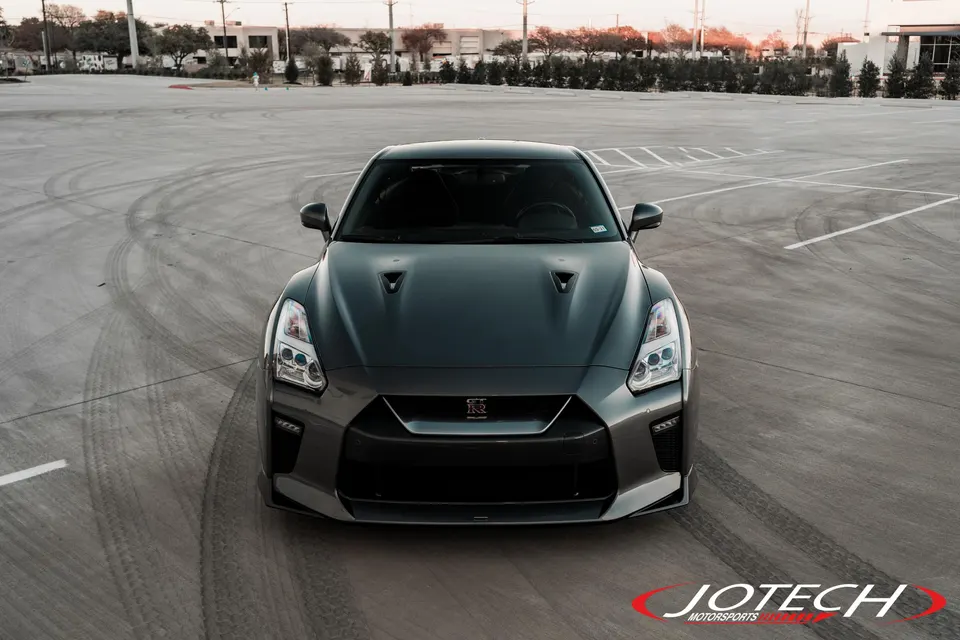 Front view of a dark grey Nissan GT-R R35 parked in a large concrete lot with tire marks.