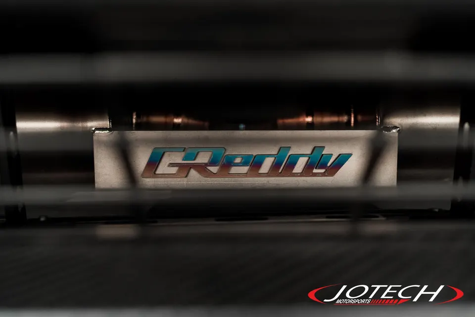 A close-up shot of a GReddy logo on a performance exhaust component, showcasing the metallic finish.