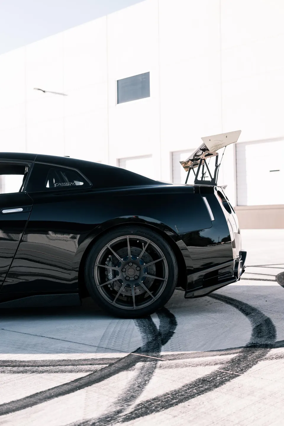 Black Nissan GT-R with a large aftermarket wing, performance wheels, and carbon fiber rear diffuser parked on concrete with tire marks.