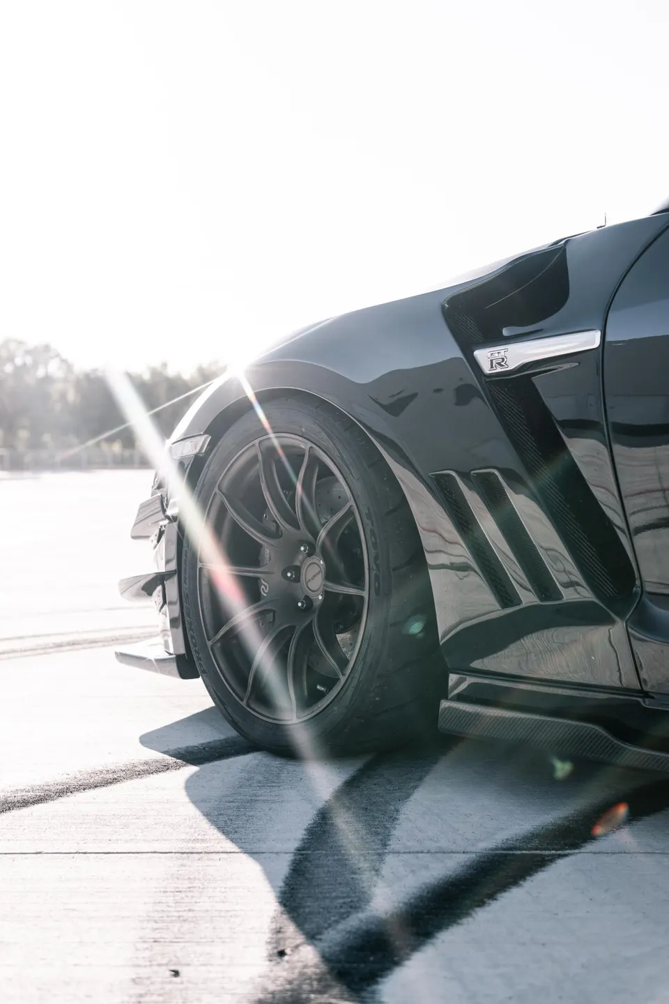 A black Nissan GT-R's front wheel and fender with carbon fiber accents, bathed in bright sunlight with a lens flare.