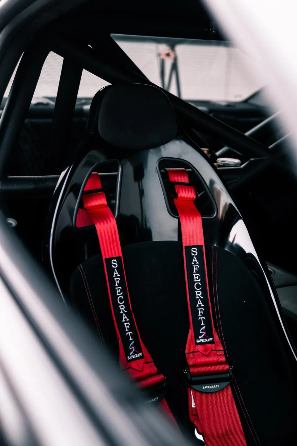 Close-up of a racing seat with red Safecraft harnesses and a roll cage in a performance car interior.