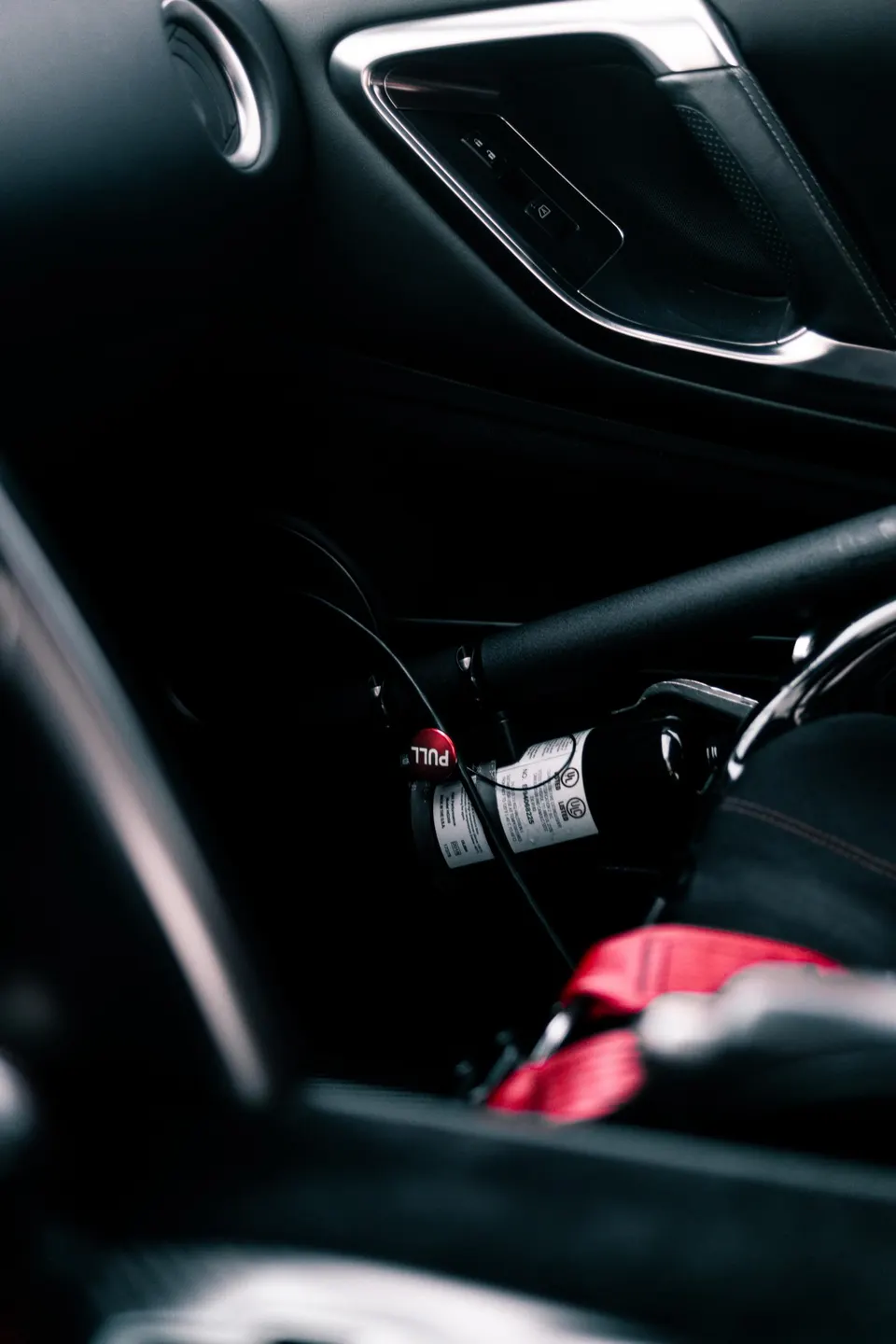 Close-up of a car's interior showing a fire extinguisher, roll bar, and red racing harness.