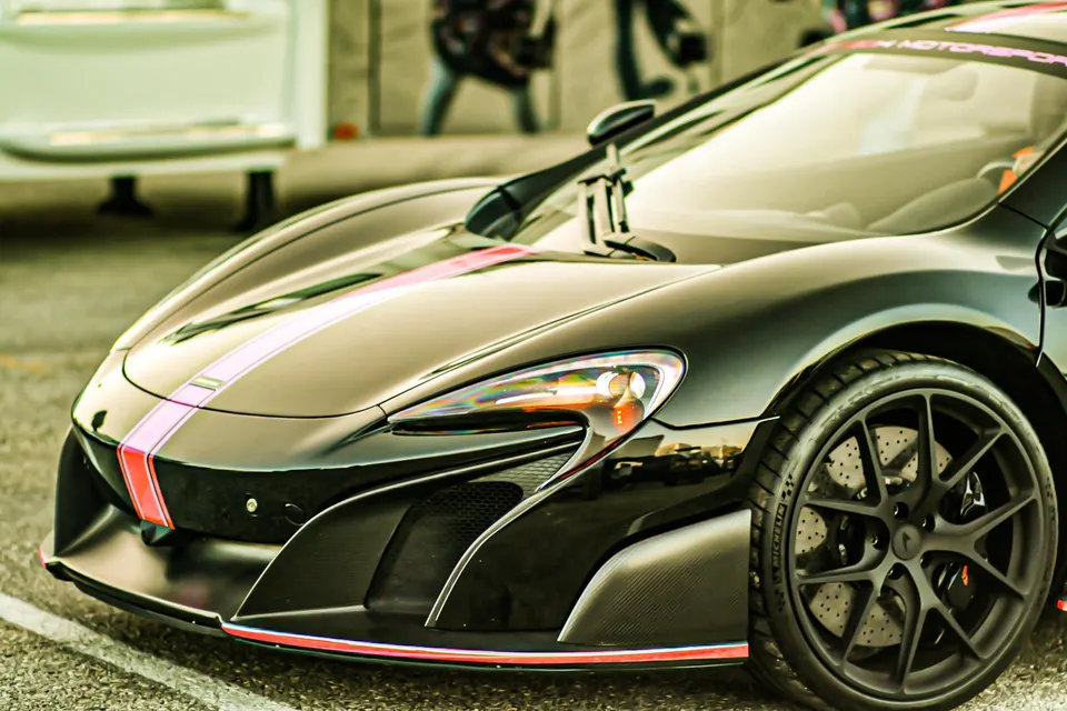 Front quarter view of a black McLaren supercar with pink and red racing stripes, showcasing its aerodynamic design and black wheels.