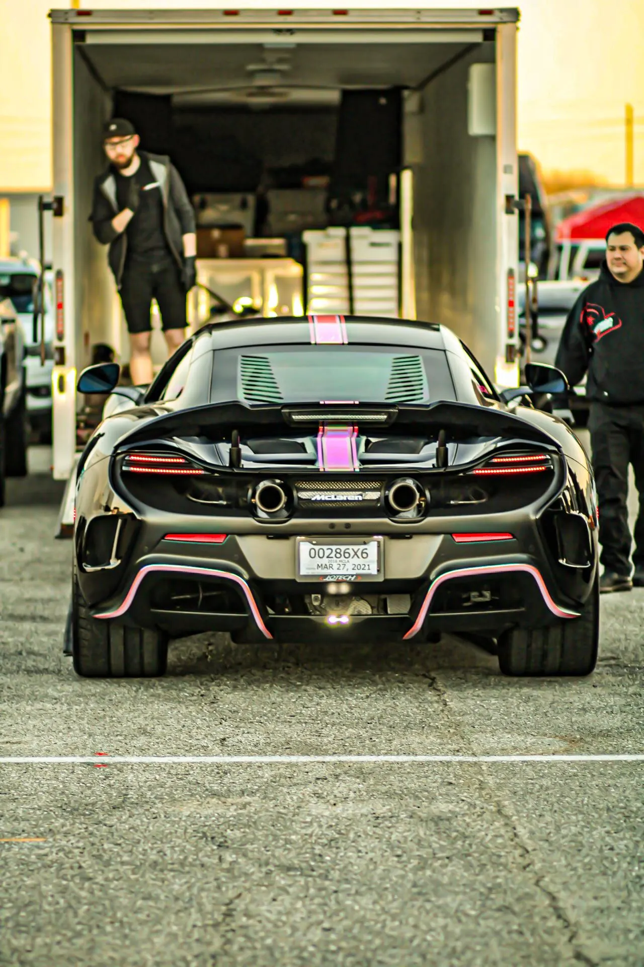 Black McLaren supercar with pink accents and dual exhaust pipes, parked at an event with a trailer in the background.