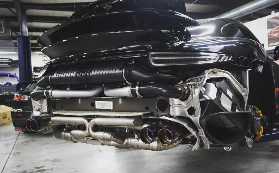 Rear view of a black Porsche 911 in a shop, showcasing a custom titanium exhaust system and intercooler setup.