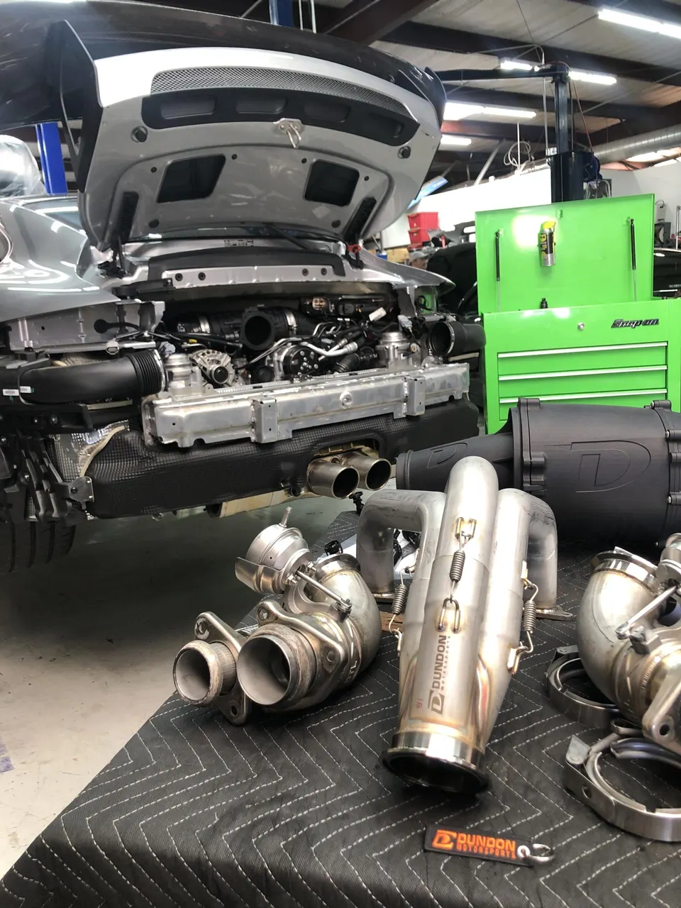 Porsche 911 with engine bay open, displaying turbochargers, exhaust system, and performance parts on a table in a shop.