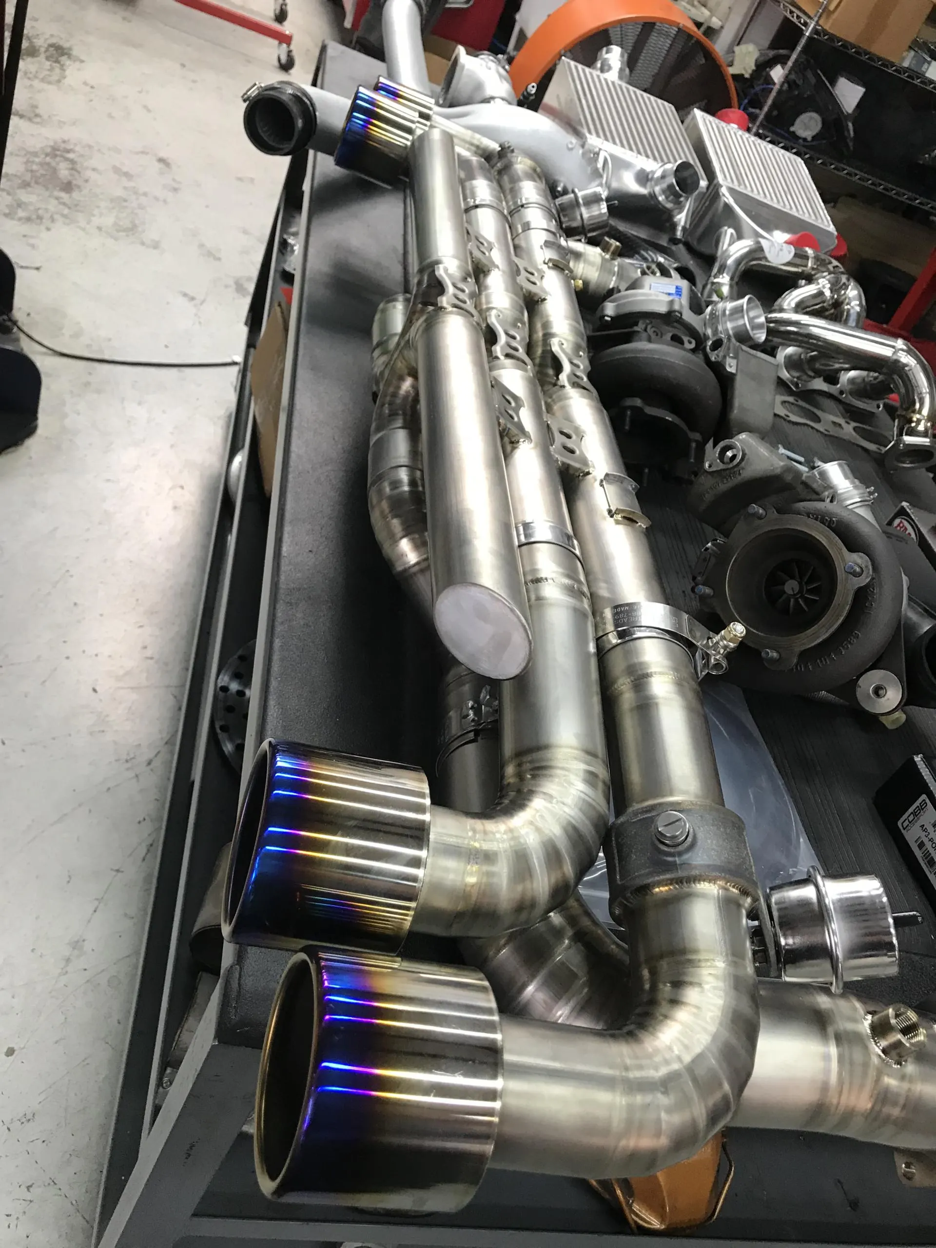 High-performance titanium exhaust system with burnt tips, turbochargers, and intercoolers laid out in a tuning shop.