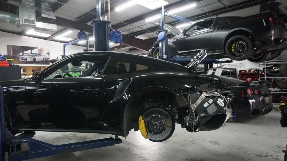 Black Porsche 911 and Nissan GT-R on lifts in a performance tuning shop, with the Porsche's rear bumper removed and brakes visible.