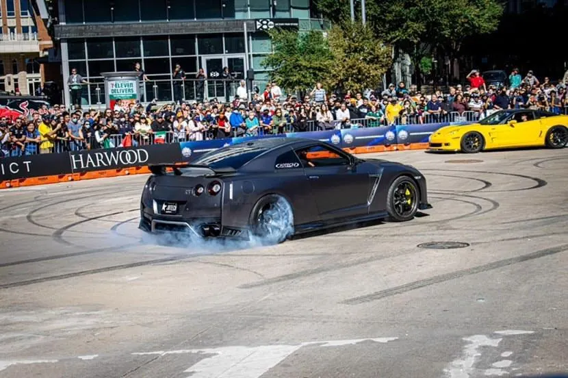 A grey Nissan GT-R performs a burnout, generating smoke from its tires, in front of a large crowd at an outdoor car event. A yellow Corvette is also visible in 