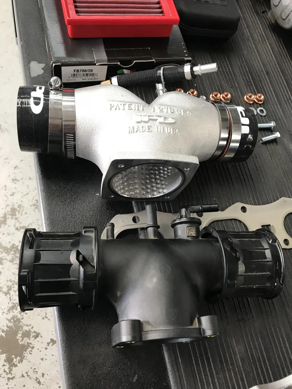 Aftermarket SPD intake manifold and throttle body assembly next to a stock unit, with an air filter in the background.