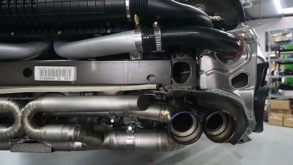 A close-up view of a performance titanium exhaust system with carbon fiber tips and intake piping on a car in a shop.