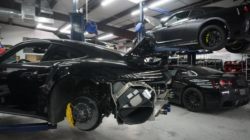Black Porsche 911 with rear bumper removed exposing exhaust and turbo components, next to two Nissan GT-Rs in a performance tuning shop.