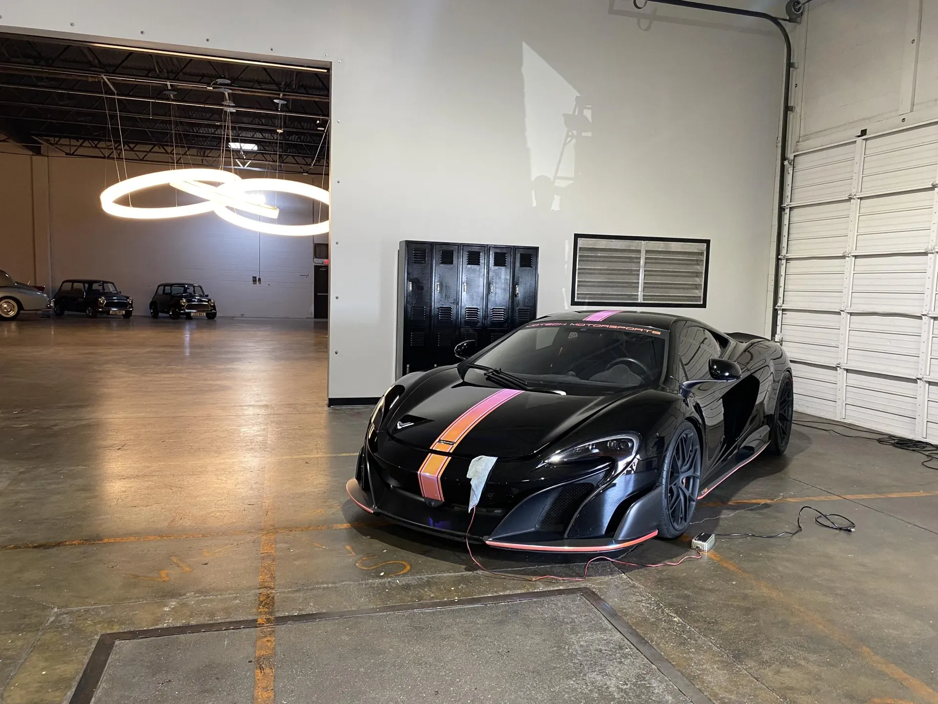 A black McLaren with pink and orange racing stripes parked in a garage with other cars in the background.