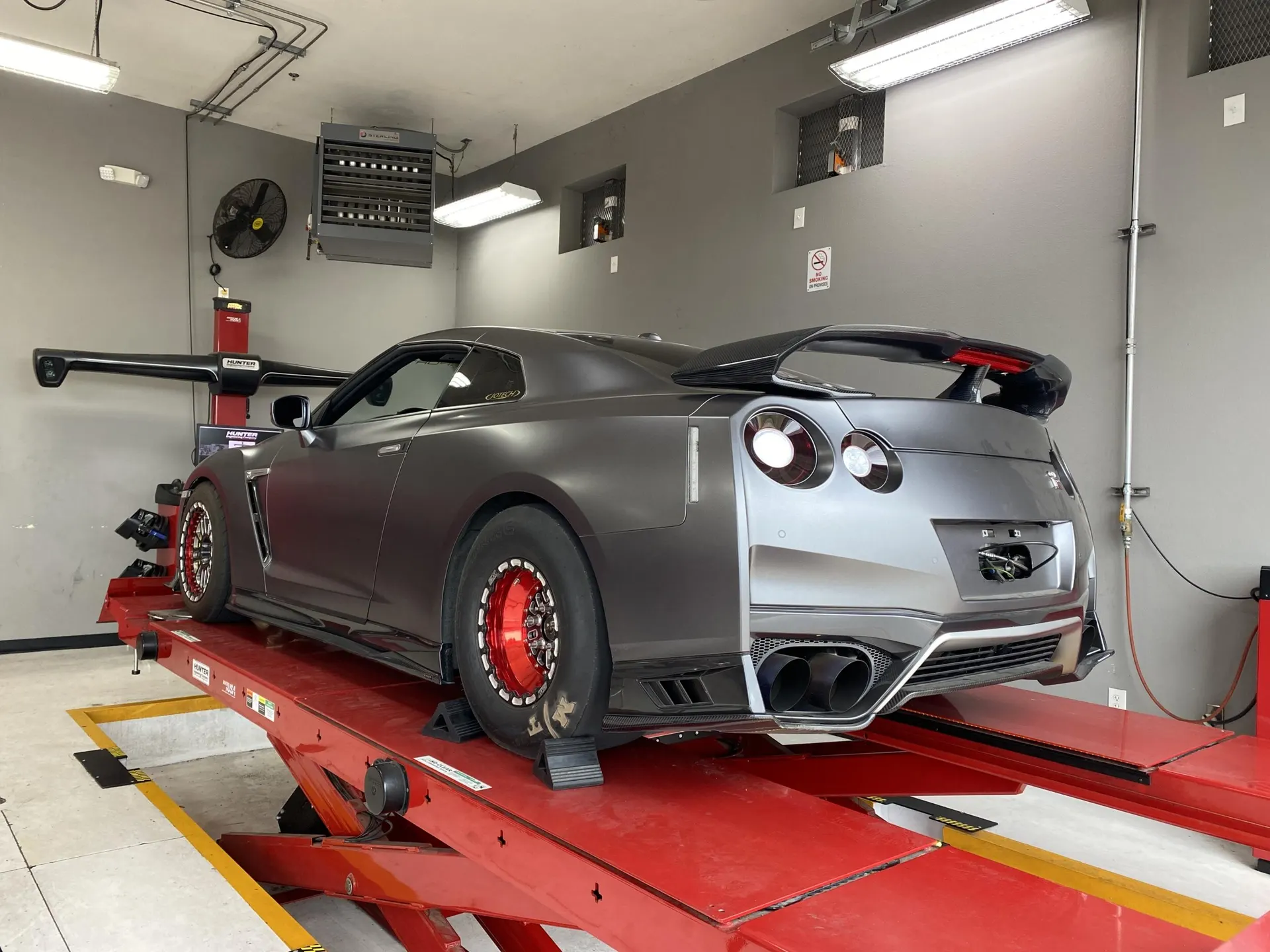 A matte grey Nissan GT-R on a red shop lift with drag wheels and a large rear wing, ready for tuning or alignment.