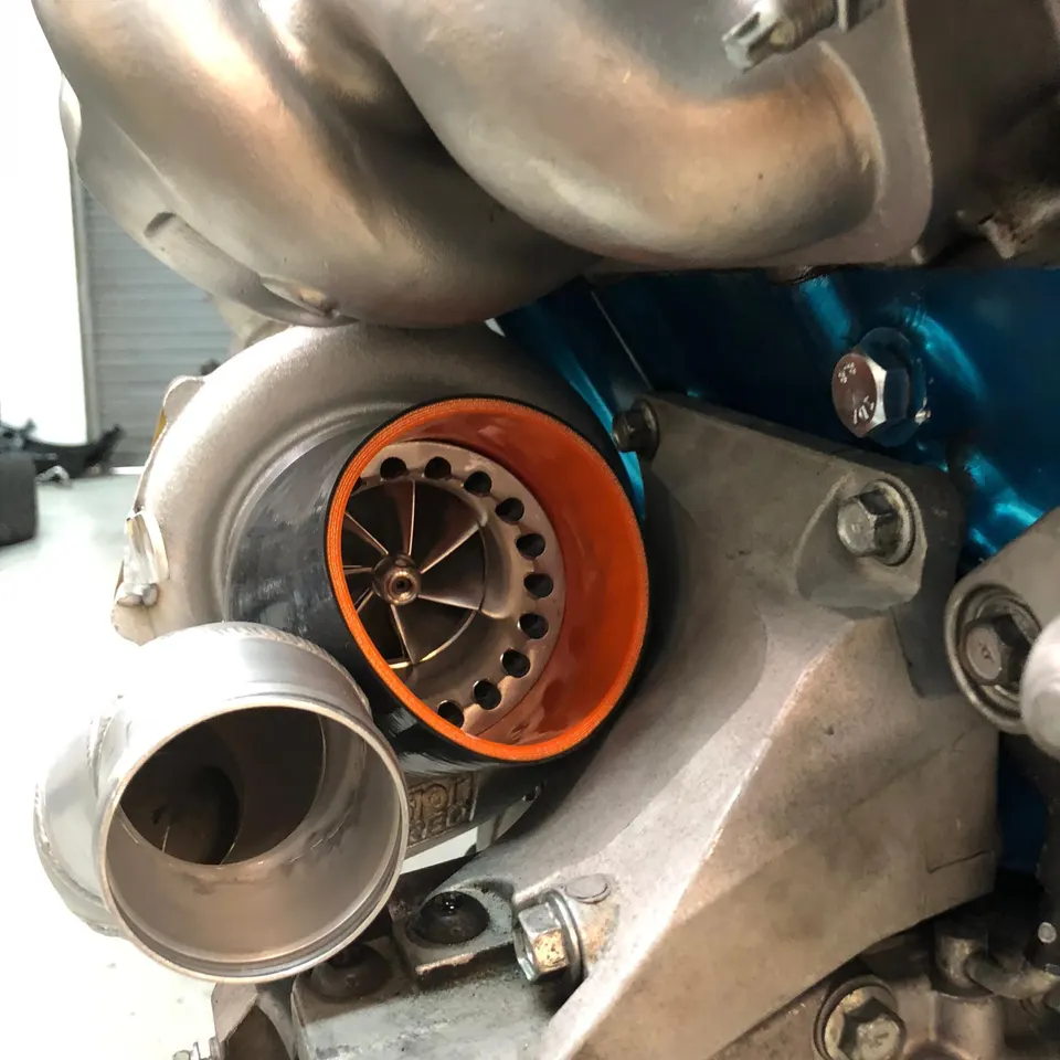A close-up view of a performance turbocharger with an orange silicone coupler and visible compressor wheel in an engine bay.