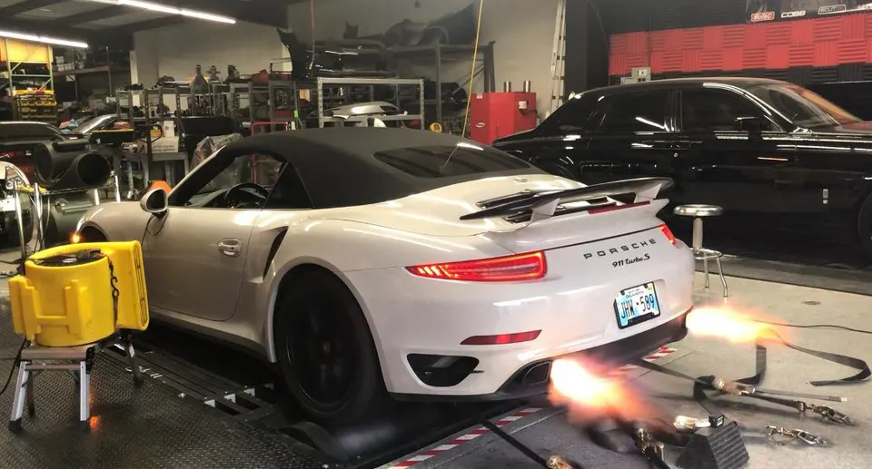 A white Porsche 911 Turbo S convertible on a dyno with flames shooting from the exhaust pipes during a tuning session.