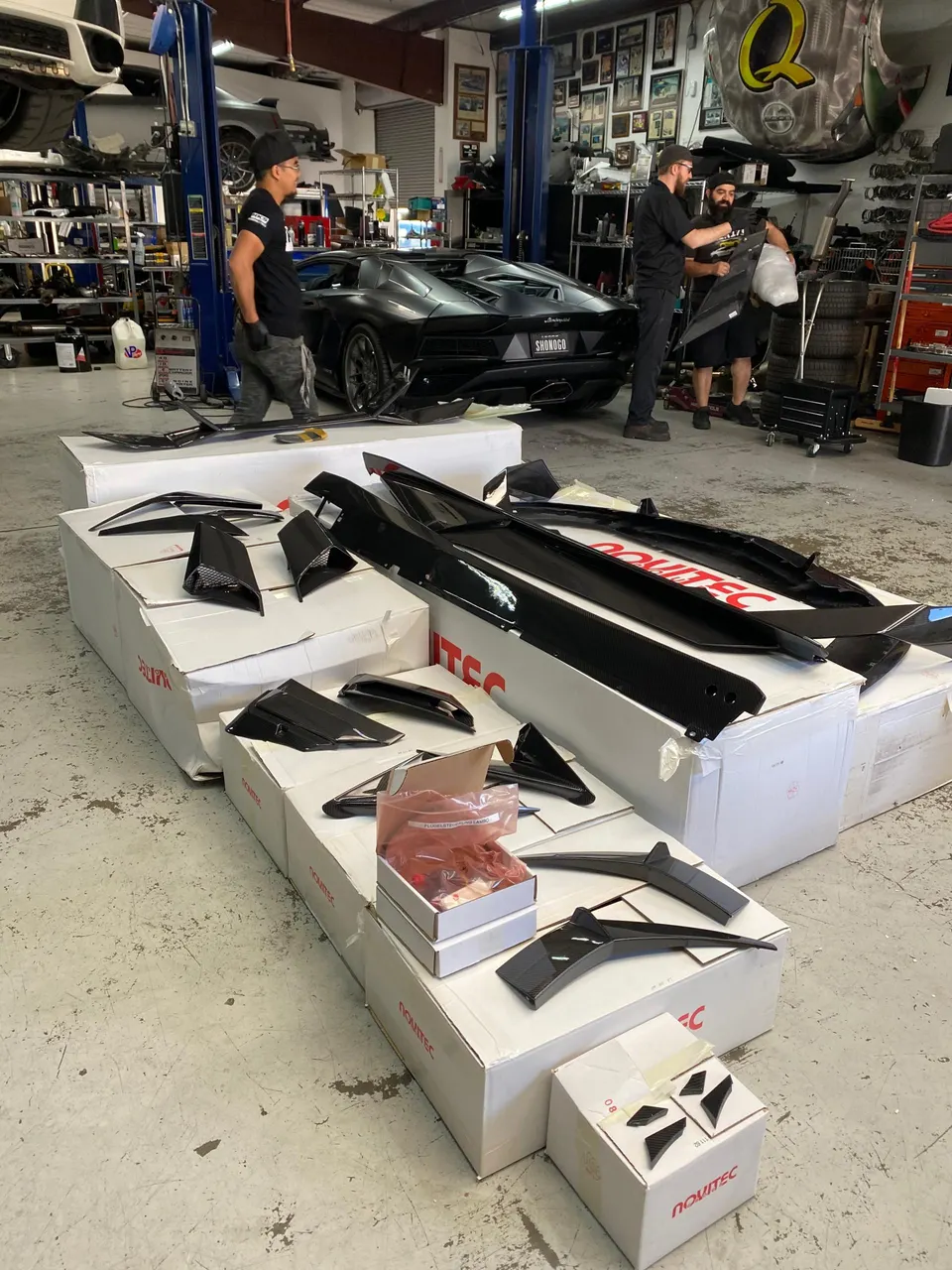 Mechanics working in a car shop with a black Lamborghini and various Novitec carbon fiber aero parts laid out on boxes.