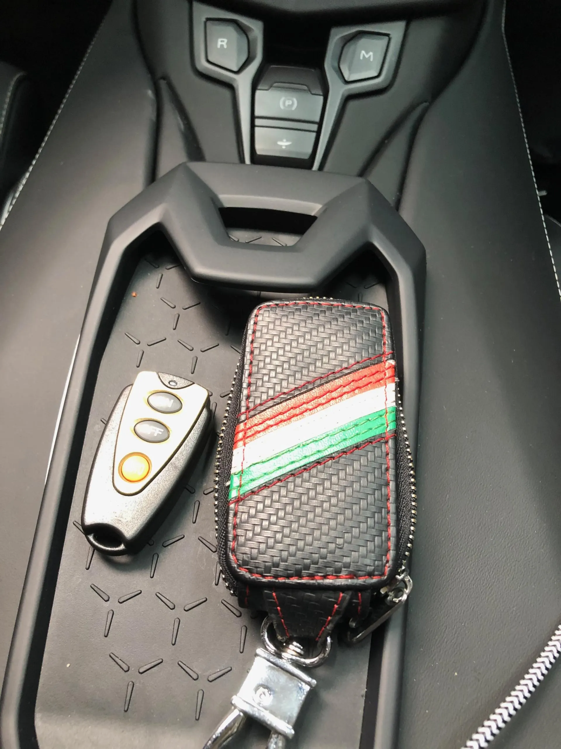 Close-up of car keys, including a key fob and a carbon fiber pattern key case with Italian flag colors, on a luxury car's center console.