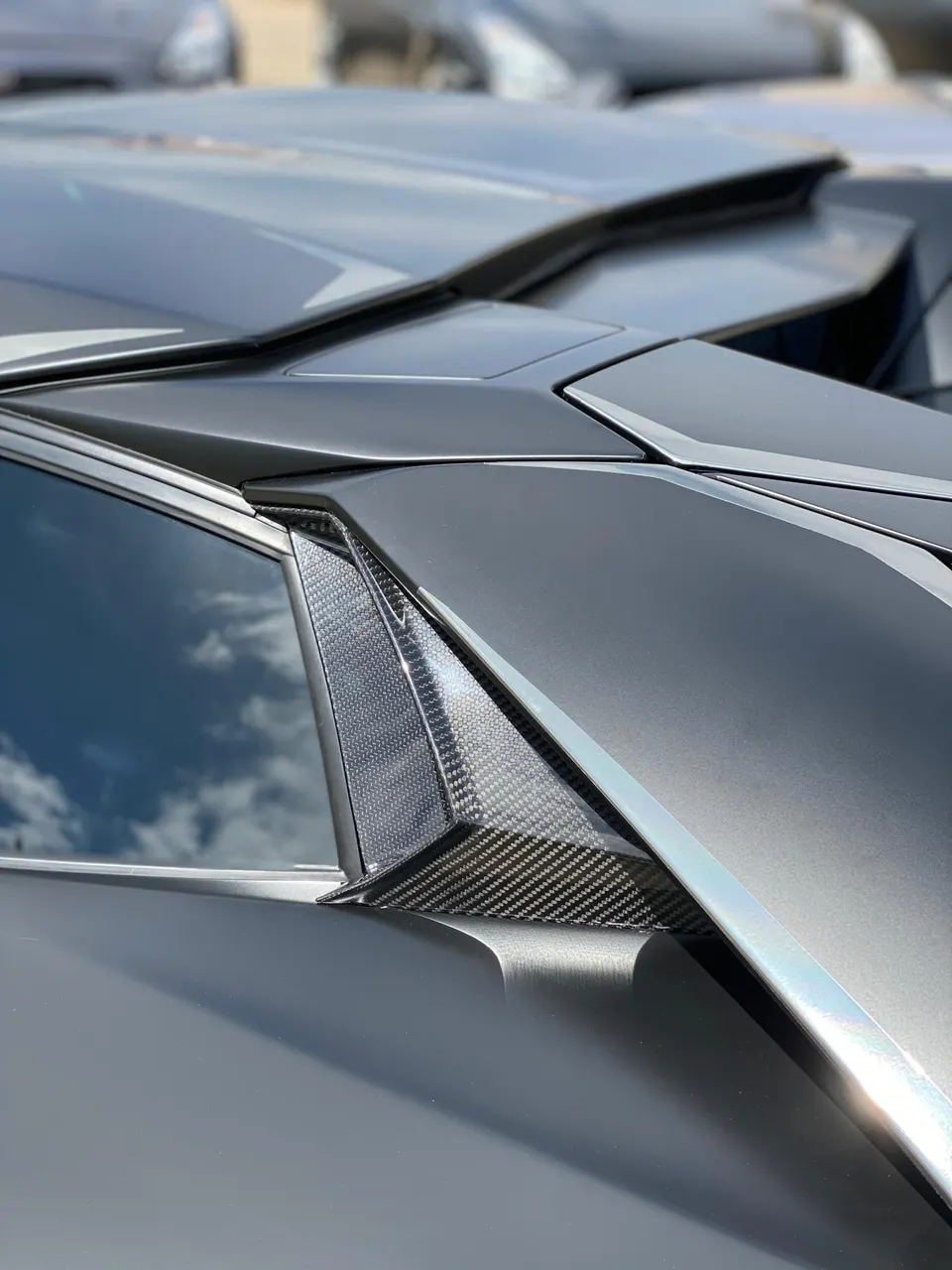 Close-up detail of a matte grey supercar's rear quarter panel with carbon fiber trim and sky reflection.