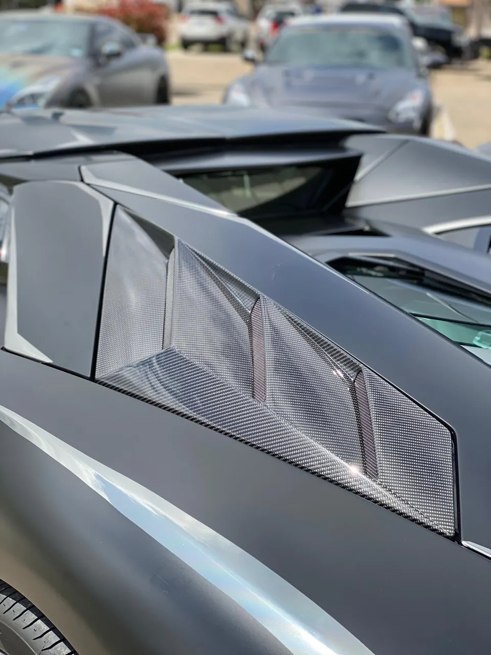 Close-up of a matte black Lamborghini's carbon fiber engine cover and vents, with other cars blurred in the background.