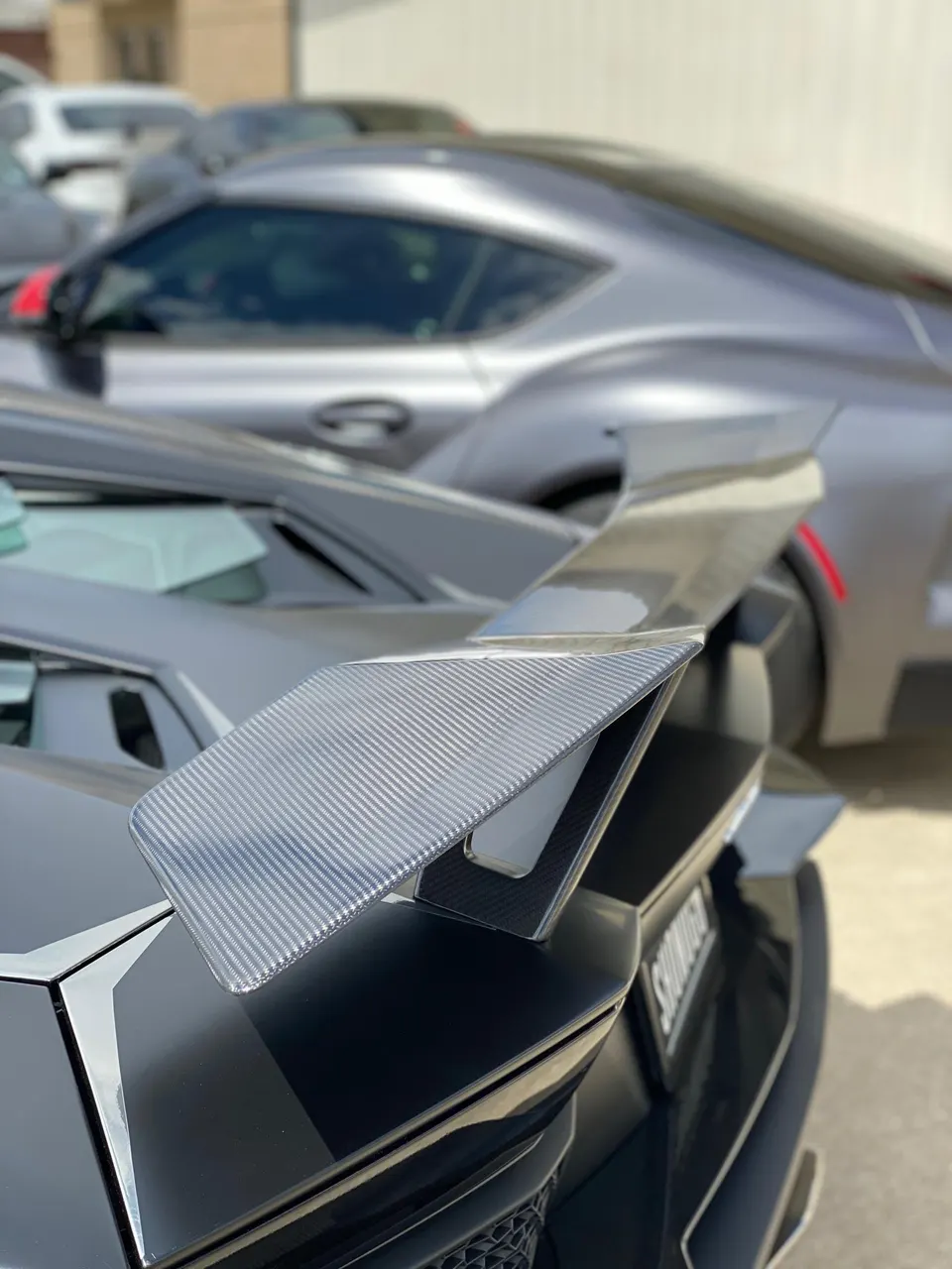 A close-up of a carbon fiber rear wing on a grey sports car, with a blurred Toyota Supra in the background.