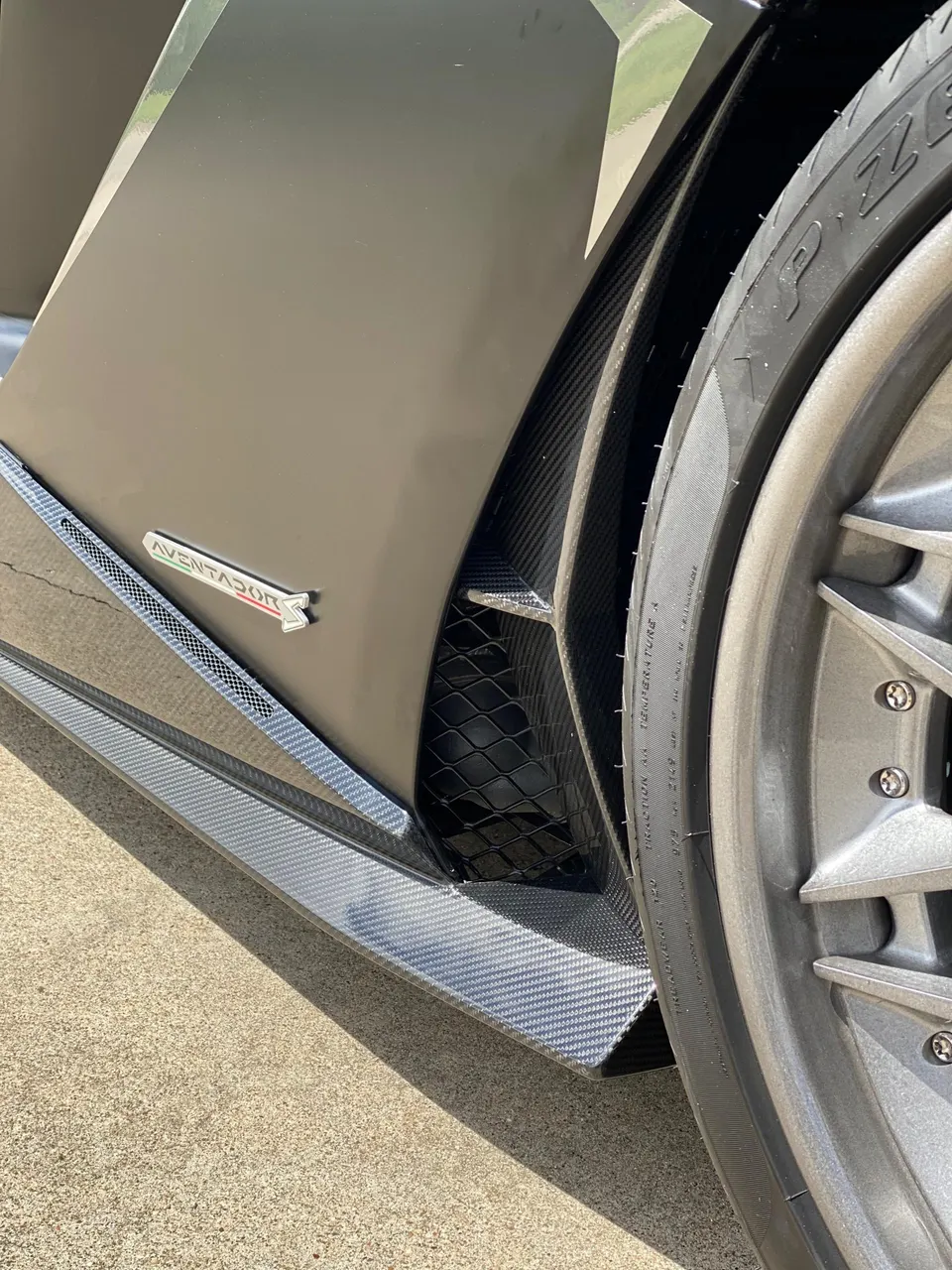 A close-up detail shot of a grey Lamborghini Aventador's carbon fiber side skirt, fender, and front wheel.