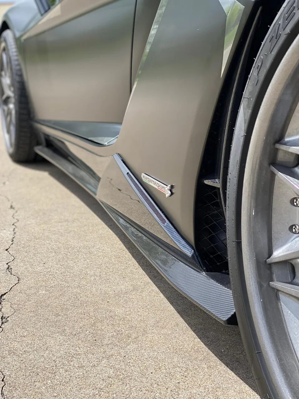 Close-up of a matte green Lamborghini Aventador's carbon fiber side skirt and badge on concrete.