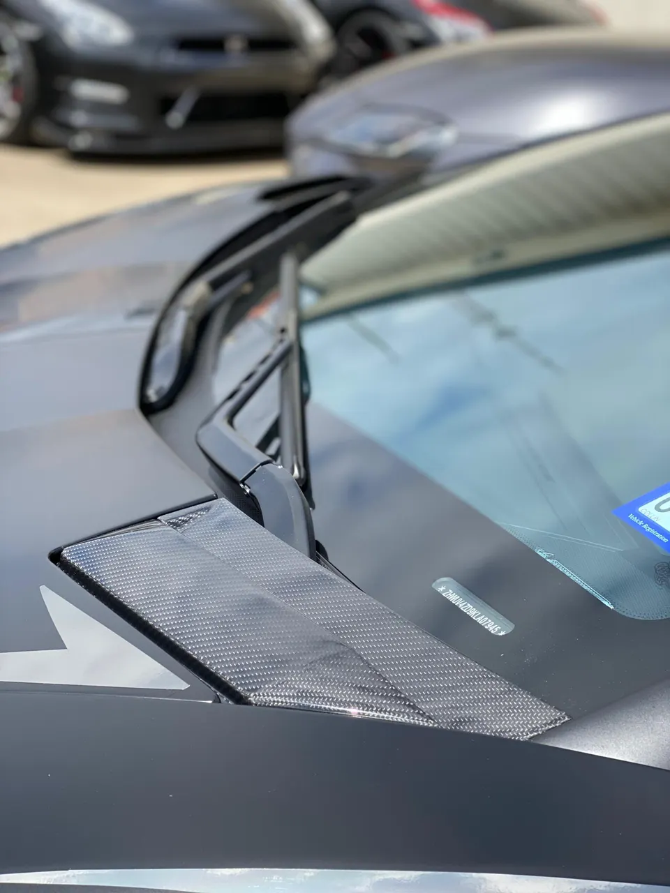 Close-up of a carbon fiber cowl vent on a performance car, with a Nissan GT-R blurred in the background.