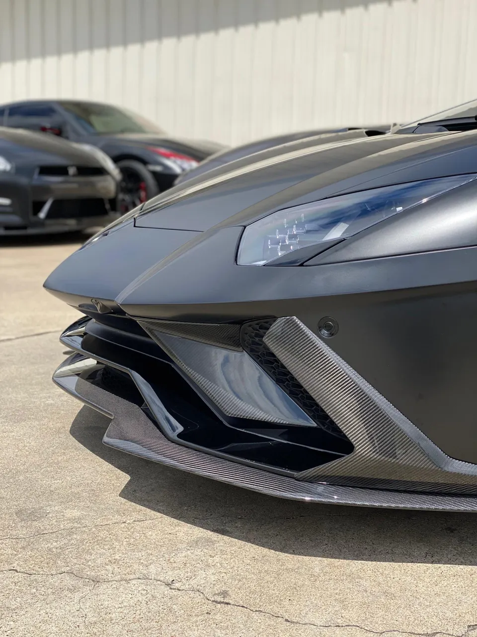 Matte black Lamborghini Aventador with carbon fiber front splitter and aero elements, parked outdoors with other sports cars.