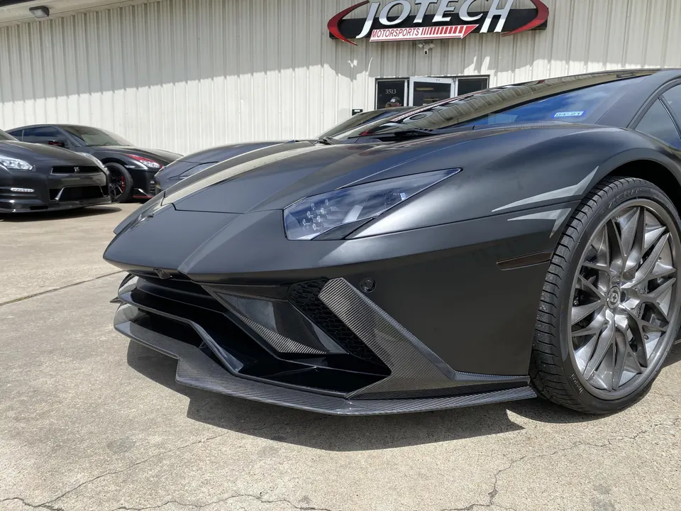 Matte grey Lamborghini Aventador with carbon fiber front splitter parked outside Jotech Motorsports, with Nissan GT-Rs in the background.