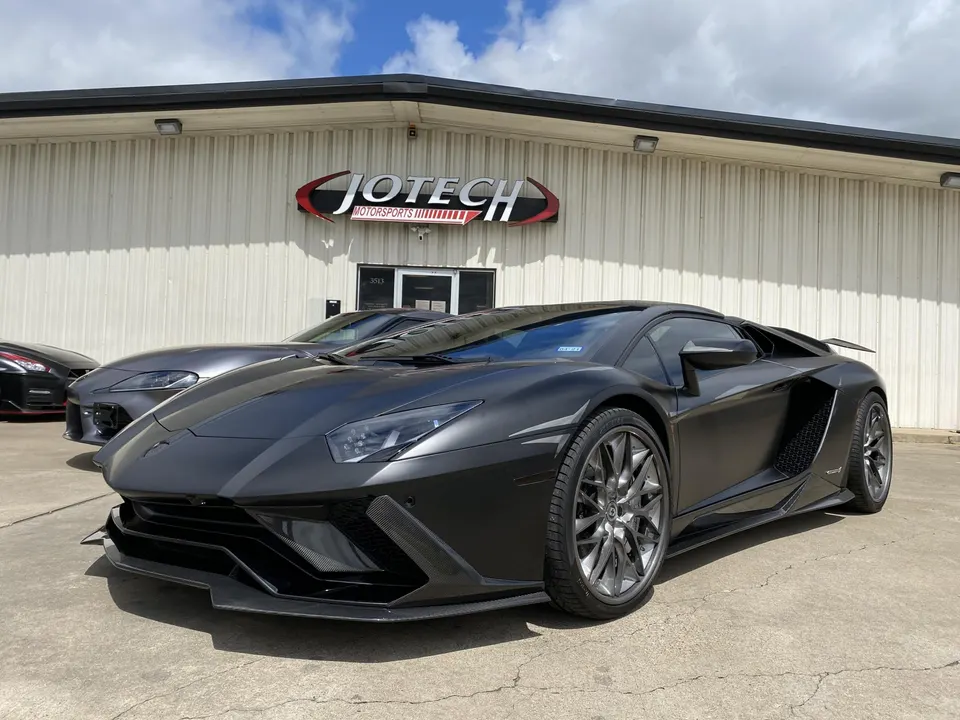 Matte black Lamborghini Aventador parked outside Jotech Motorsports with a Toyota Supra and Nissan GT-R in the background.